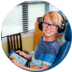 A smiling child with glasses and headphones holds a tablet, seated in a colorful room with a cheerful atmosphere. Bright window blinds in the background.