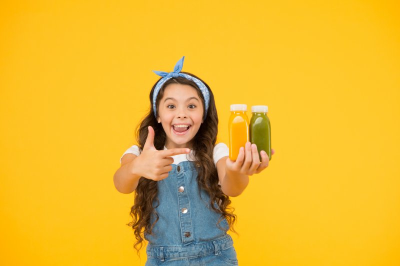 a young girl wearing a denim romper holding two bottles of juice and smiling