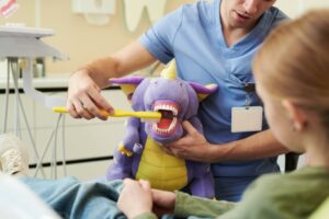 A pediatric dentist demonstrating tooth brushing using a large toothbrush on a purple dragon puppet for a young patient