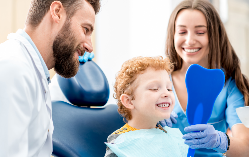 Child at dentist smiling into mirror that the dental assistant is holding while the dentist also looks on and smiles